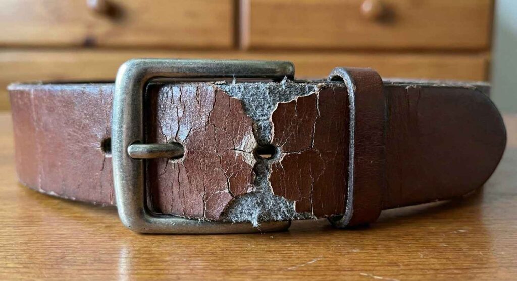 A close-up photograph of a cheap, cracked 'genuine leather' belt showing the peeled surface and fibrous interior near the buckle