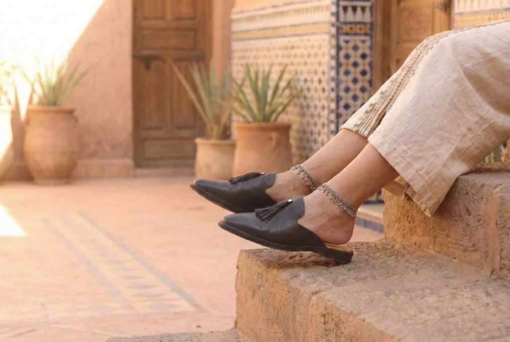 Side view of black leather Malika Babouche slippers with tassel, worn by a woman sitting on steps in a riad