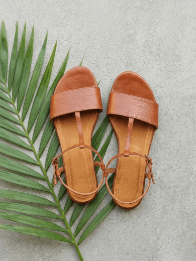 Flat lay of The Tanalt tan leather T-strap sandals on stone with green palm frond